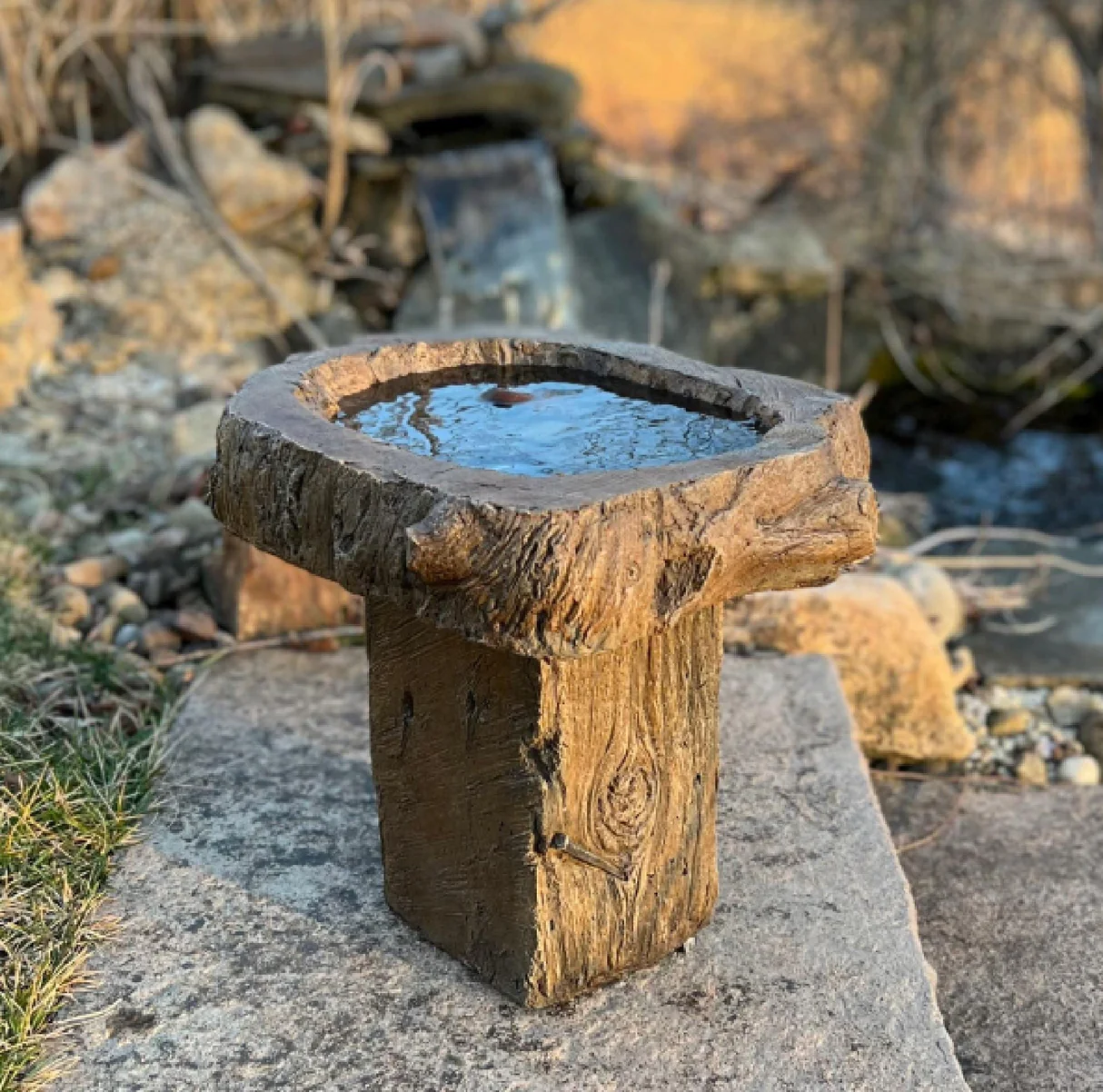 Rustic handmade bird bath designed to resemble natural wood, placed on a stone slab beside a small garden stream and rocks.
