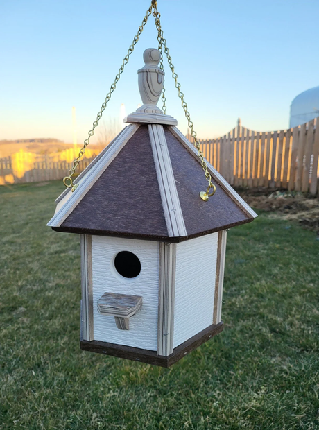 Hanging hexagonal white birdhouse with small perch, brown roof, and gold chains against a rural backdrop