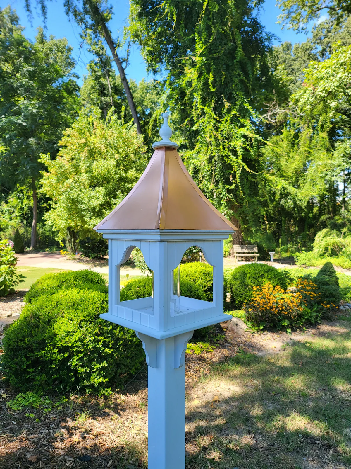 Tall white post-mounted bird feeder with scalloped arches and curved copper roof in a landscaped garden