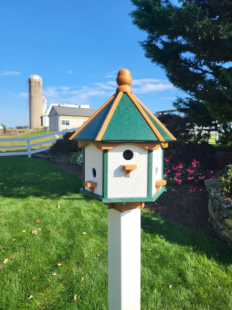 "Handcrafted bird feeder house with a green and natural wood-tone roof, made from weather-resistant poly lumber, mounted on a white post in a scenic farm setting."