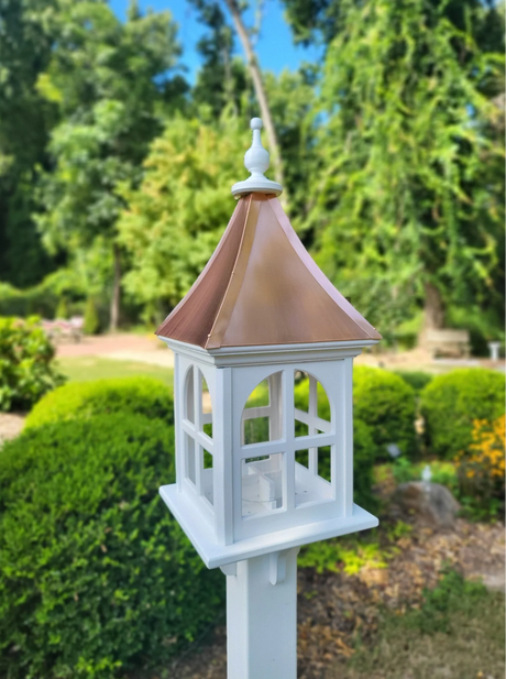 Elegant copper bird feeder with a white wooden base and handcrafted copper roof, enhancing a garden space with durability and style—available at BetterCrafter.com.