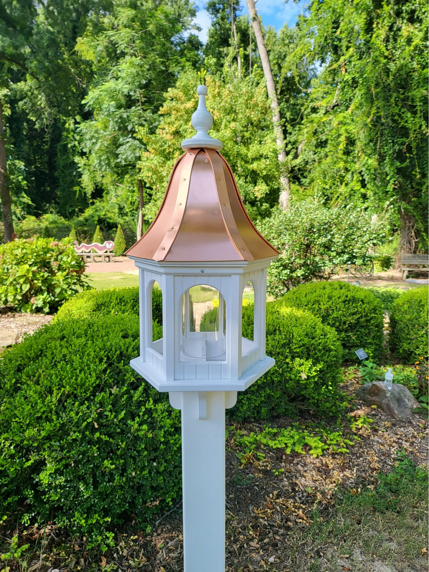 Beautiful white bird feeder with a copper roof mounted on a white post, set in a lush garden with vibrant greenery and a serene outdoor setting.